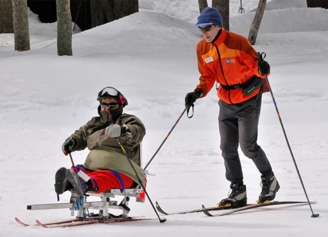 Adaptive Nordic skiing participant using a sit-ski with instructor assistance on snowy trails.