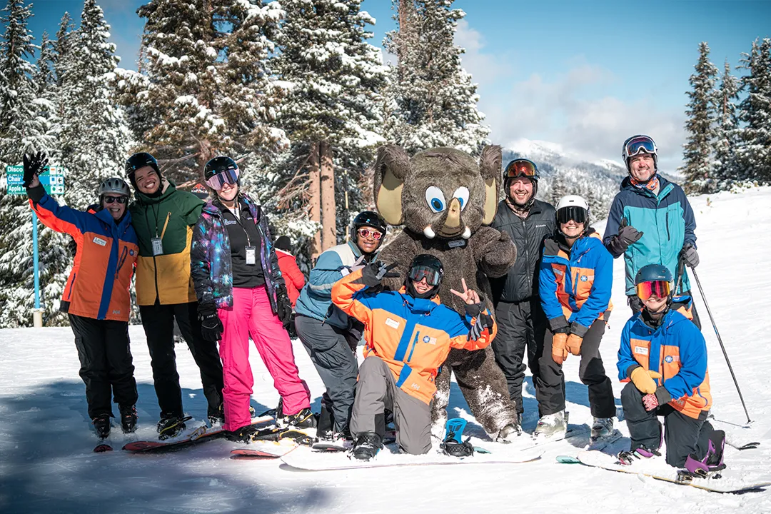 Group of Access Mammoth snowboarders and instructors posing at the top of the mountain with the Mammoth Mountain mascot.