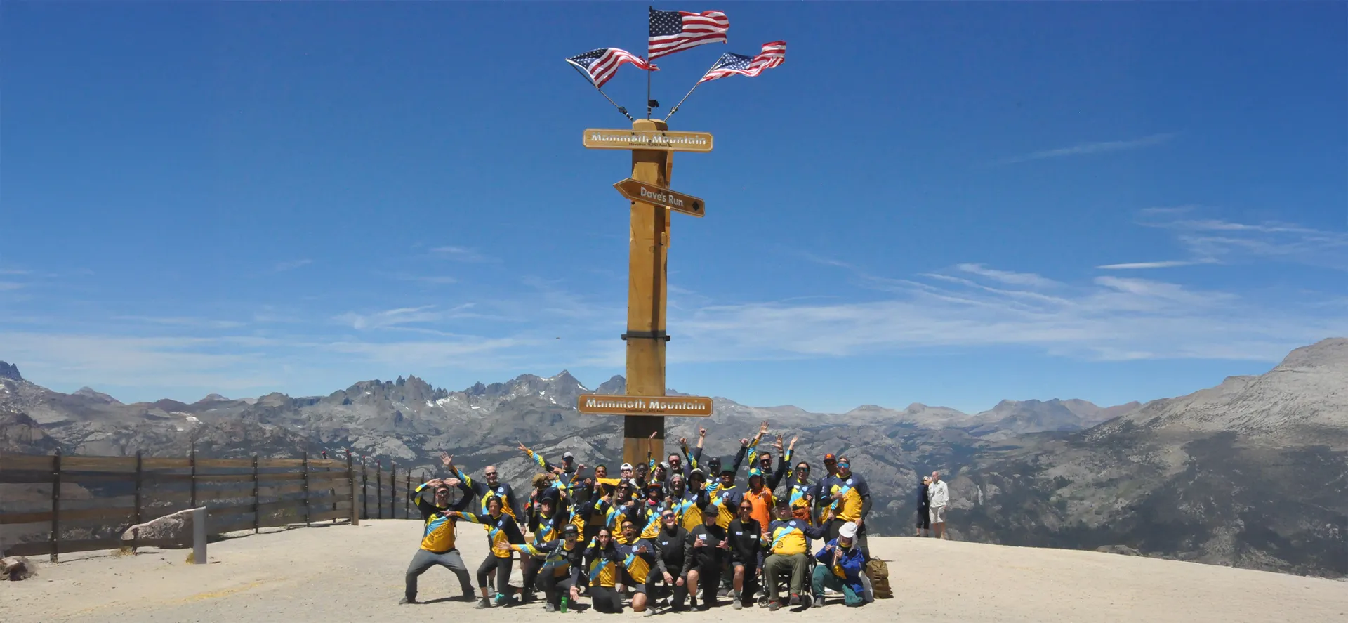 Group of adaptive athletes and supporters posing at Mammoth Mountain summit under American flags with panoramic Sierra Nevada views.