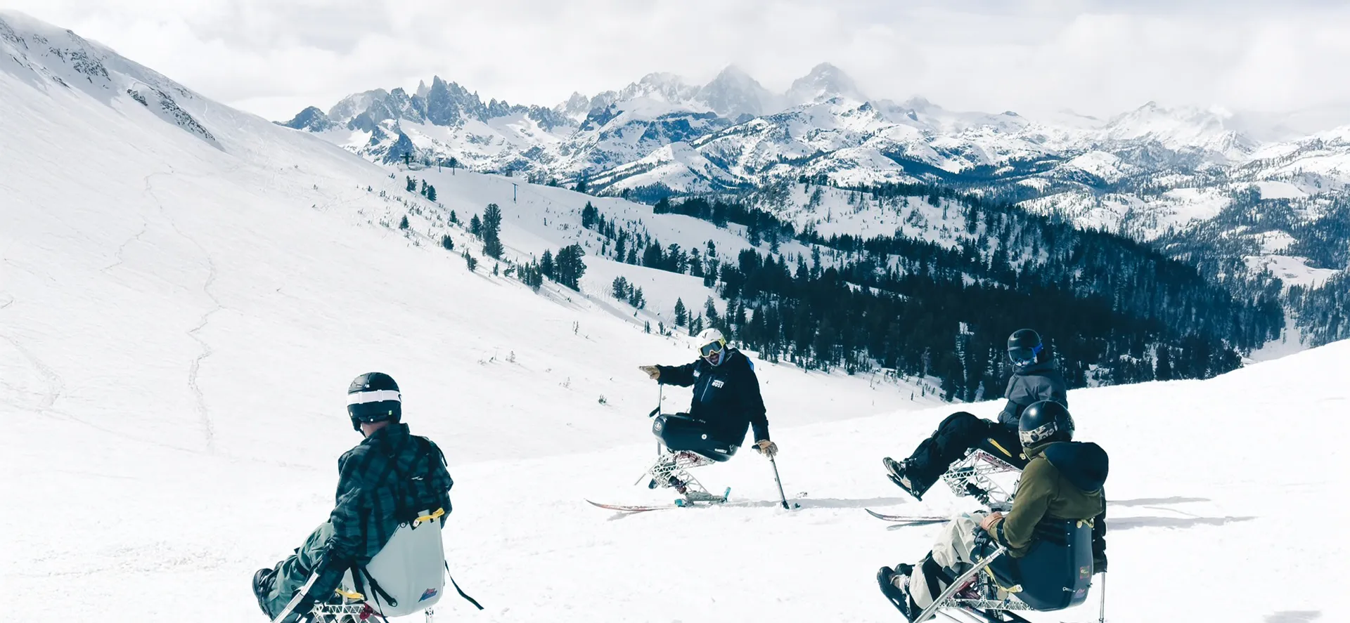 Group of adaptive skiers using mono-skis on a snowy mountain slope with scenic alpine peaks in the background.