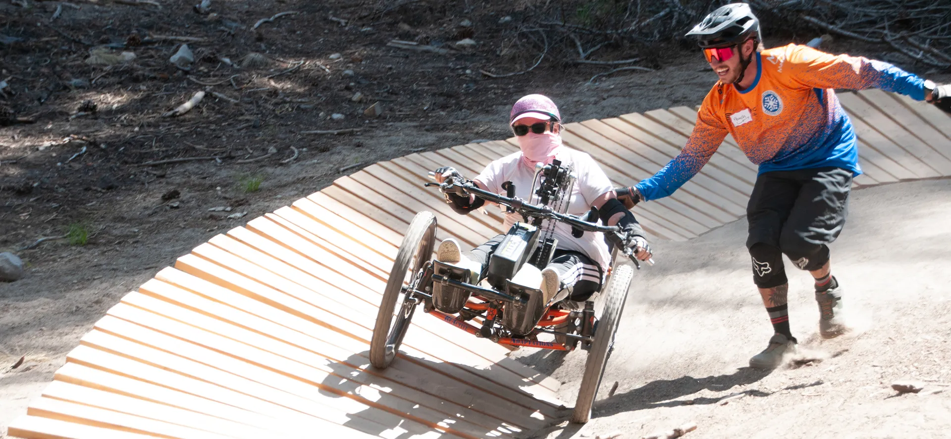 Adaptive mountain biking rider on a three-wheel off-road bike assisted by a guide on a dirt trail during an outdoor camp.